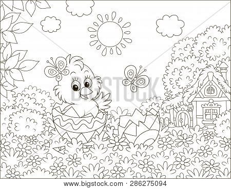 Little Chick Peeking Out Of A Decorated Easter Egg On Grass Among Flowers On A Lawn Near A Small Hut