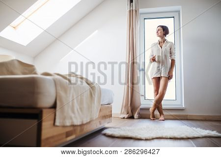 A Young Woman With Night Shirt Standing By The Window In The Morning.