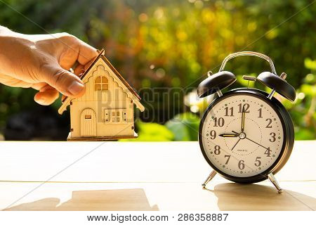 Man Catch Home Model And The Alarm Clock Stack On Wood Table And Sunset Background In The Public Par