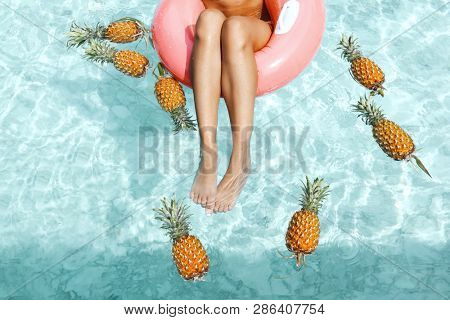Girl relaxing on float ring in pool with pineapples in blue clear water, photo of legs closeup. Tropical fruit diet. Summer holiday idyllic concept top view.