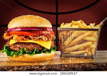 Cheeseburger With Bacon And A Basket Of French Fries. Basketball Ball Image In Background.