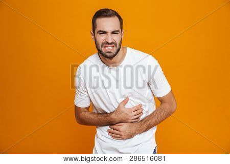Image of caucasian man 30s in t-shirt bending and touching his belly because of pain while isolated over yellow background