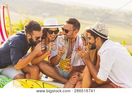 Group Of Friends At A Poolside Summer Party, Sitting At The Edge Of A Swimming Pool, Drinking A Pine