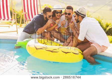 Group Of Friends At A Poolside Summer Party, Sitting At The Edge Of A Swimming Pool, Drinking A Pine