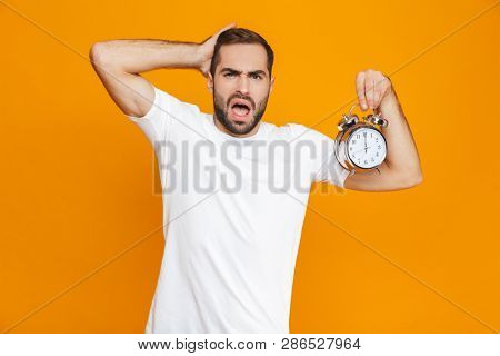 Photo of uptight man 30s in casual wear holding alarm clock isolated over yellow background