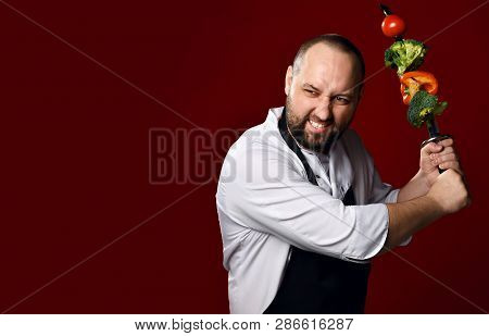 Bearded Professional Chef In White Skirt And Apron Going To Fight With Skewer With Vegetables On It 