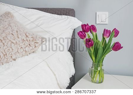 Purple Tulips Standing On A Nightstand In The Modern White Bedroom Near The Bed.