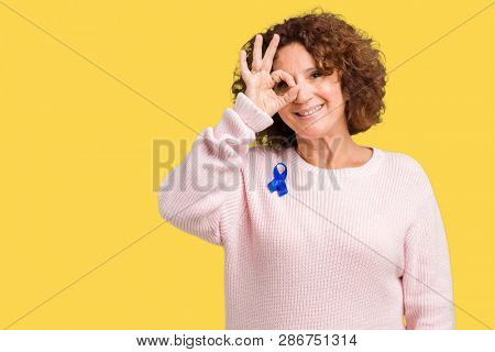 Middle ager senior woman wearing changeable blue color ribbon awareness over isolated background doing ok gesture with hand smiling, eye looking through fingers with happy face.