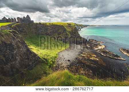 Northern Ireland shoreline with Dunluce castle in the distance. Picturesque grass covered lowland and the steep cliffs under the grey cloudy sky. Amazing Irish bay before the rain. Wild virgin nature.