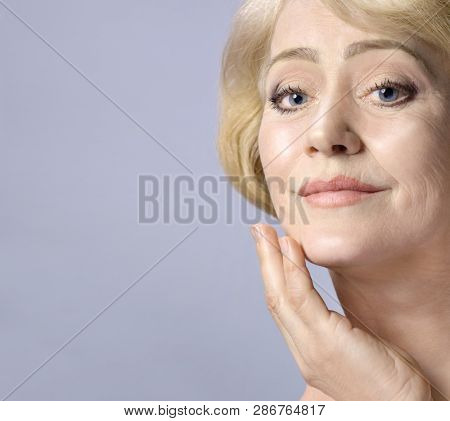 Beautiful Matured Woman Tauching her Face with hand. cheerful caucasian smiling blond. Studio shot. Looking off camera. Facecare. Grey background. 50 . Skin care concept