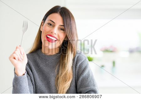 Young beautiful woman holding silver fork at home with a happy face standing and smiling with a confident smile showing teeth