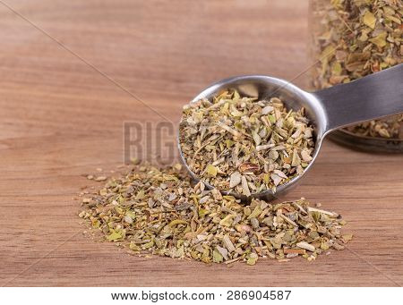 Closeup Of A Measuring Spoon Of Chopped Oregan Spilled Onto A Wooden Surface And A Jar Of Herbs In B