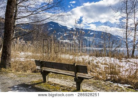 Old Wooden Bench In The Sun Facing Out To Snow Covered Path And Lake With Mountains With Snow In The
