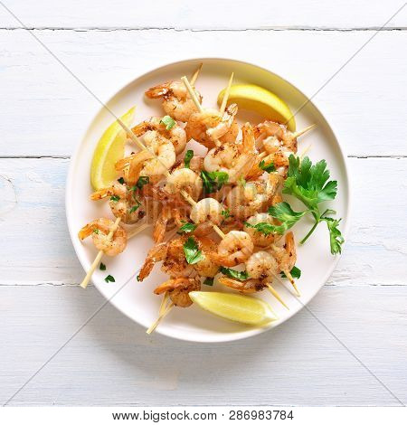 Grilled Shrimp Skewers. Prawns Skewers With Greens, Spices And Lemon On White Plate Over Wooden Tabl