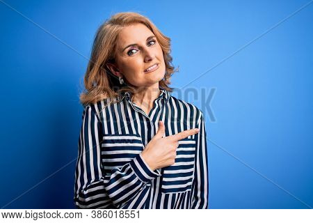 Middle age beautiful blonde woman wearing casual striped shirt standing over blue background cheerful with a smile of face pointing with hand and finger up to the side with happy and natural expressio