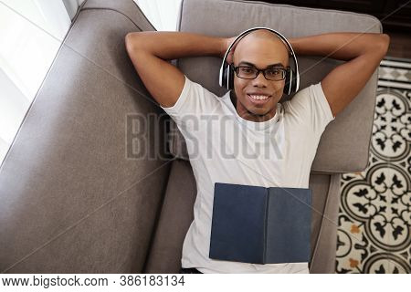 Happy Handsome Young Black Man In White T-shirt Relaxing On Sofa, Listening To Music In Headphones A