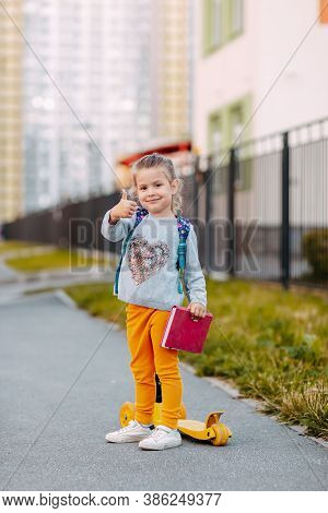 Little Girl Goes To School For The First Time. Little Student With Purple Backpack And Pink Notebook