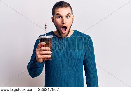 Young handsome man dirnking cola refreshment beverage scared and amazed with open mouth for surprise, disbelief face 