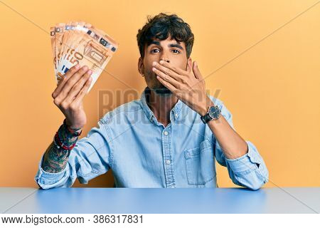 Young hispanic man holding 50 euro banknotes sitting on the table covering mouth with hand, shocked and afraid for mistake. surprised expression 