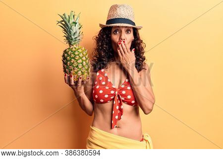 Middle age beautiful woman wearing bikini and summer hat holding pineappel covering mouth with hand, shocked and afraid for mistake. surprised expression 