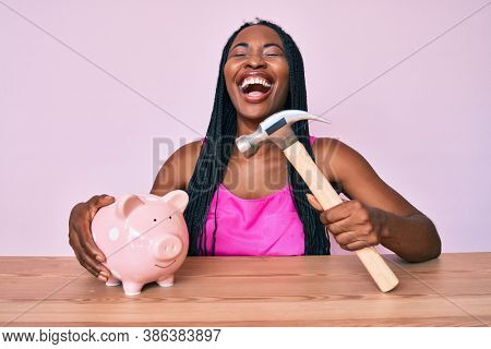 African american woman with braids holding piggy bank and hammer smiling and laughing hard out loud because funny crazy joke. 