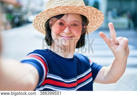 Beautiful brunette woman with down syndrome at the town on a sunny day wearing fashion heart shaped sunglasses and summer hat doing ok symbol with fingers taking a selfie picture
