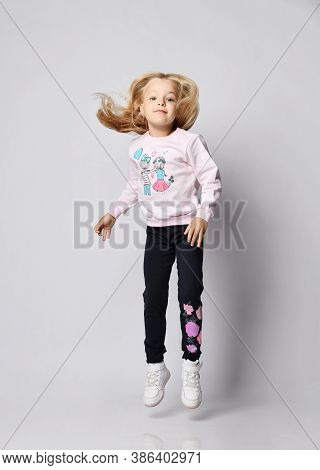 Full-growth Portrait Of Blonde Kid Girl In Gray Sweatshirt Sweater, Blue Pants And Sneakers Jumping 