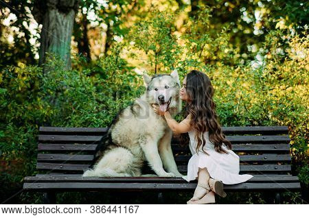 Young Woman With Innate Disorder Dwarfism Sits On Bench Next To Malamute Dog And Hugs It While Walki