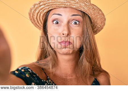 Middle age hispanic woman wearing summer hat taking a selfie puffing cheeks with funny face. mouth inflated with air, catching air. 