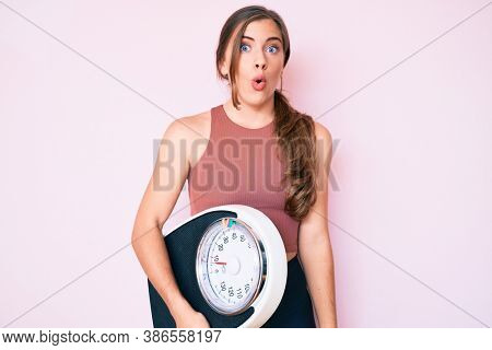 Beautiful young caucasian woman holding weight machine to balance weight loss scared and amazed with open mouth for surprise, disbelief face 