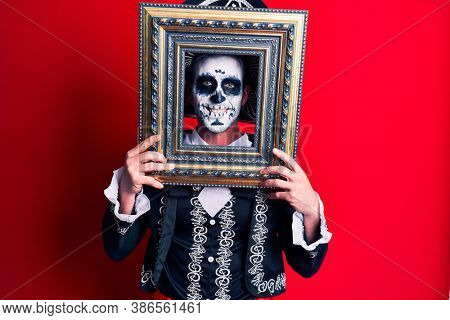 Young man wearing mexican day of the dead costume holding empty frame looking positive and happy standing and smiling with a confident smile showing teeth 
