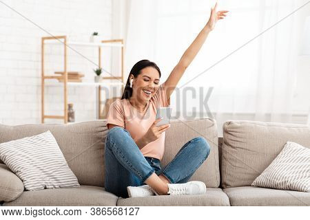 Weekend Leisure. Excited Young Woman Listening To Music In Wireless Earbuds, Sitting On Couch And Us