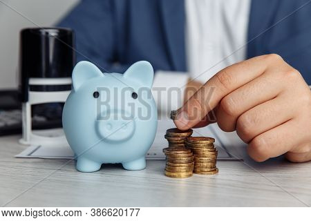 Blue Piggy Bank, Stamp And Mans Fingers With Coins On Desk In Office. Save Money And Management Fina