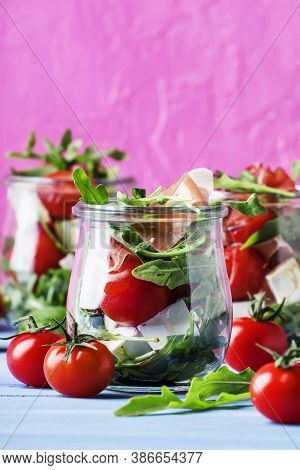 Salad With Arugula, Cheese, Cherry Tomatoes And Prosciutto In Glass Jars, Pink Blue Background, Sele