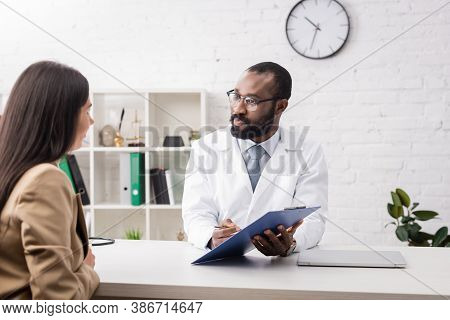 African American Doctor In Eyeglasses Holding Clipboard Near Brunette Woman