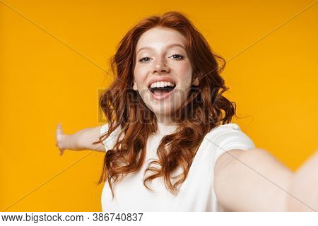 Image of redhead cheerful girl showing cpyspace and taking selfie photo isolated over yellow background