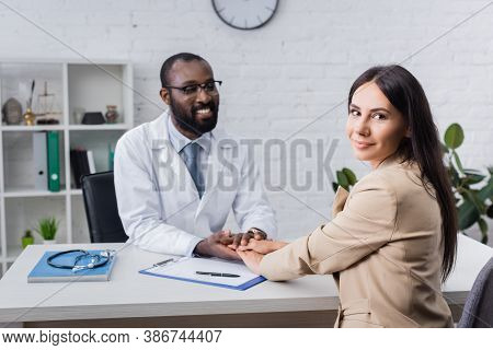 Joyful African American Doctor Touching Hands Of Pleased Patient Looking At Camera Near Clipboard Wi