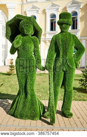 Topiary Sculpture Of Lady With Umbrella And Dressed In Flared Skirt And Jentleman With A Cane And A 