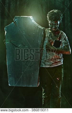 Horrible Halloween. Scary zombie boy teenager peeps out from behind the headstone and gloats in the cemetery. Copy space.
