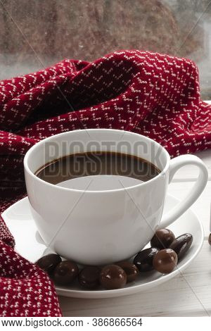 White Cup Of Coffee With Chocolates Stands On A White Window Sill Against The Window