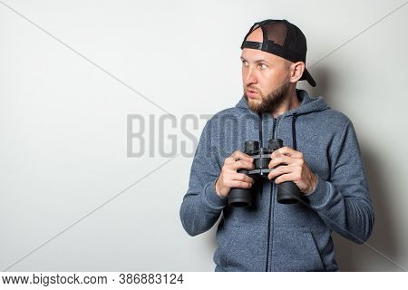 Young Man In A Hoodie And A Cap With A Serious Face Looks To The Side And Holds Binoculars On A Ligh