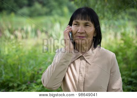 Senior Chuvash woman speaking by phone on summer green background