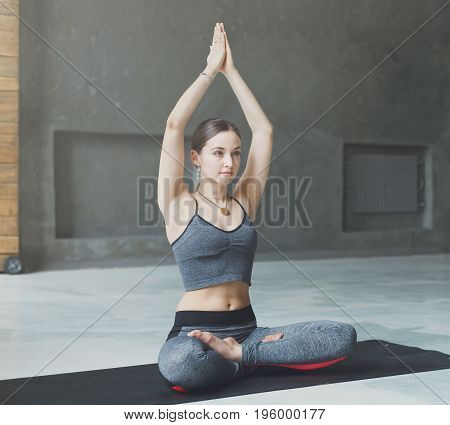 Attractive girl practicing yoga sitting in padmasana. Young woman in lotus pose doing breathing exercise on mat at sport club interior, copy space