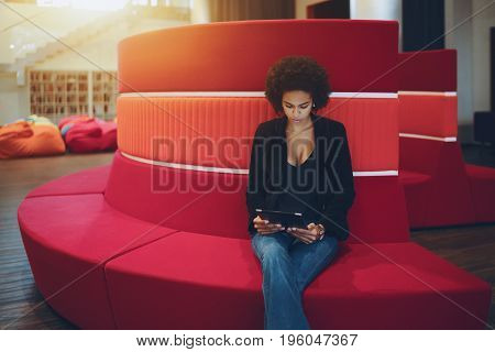 Young serious afro american girl with curly hair is sitting on red curved sofa in chillout office space and reading e-mail from her boss about future meeting with copy space place for text or logo