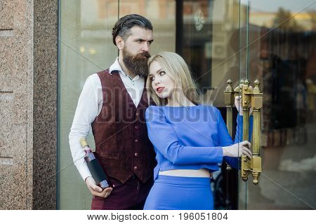 Hipster with bottle of wine hugging girl at glass door. Adorable woman in blue dress and brutal man outdoors on brick wall. Alcohol and convive. Couple in love. Unhealthy lifestyle. Bad habits