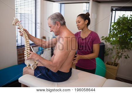 Shirtless male patient holding artificial spine while female therapist examining back at hospital ward