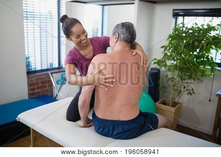 Smiling young female therapist examining back of shirtless senior male patient sitting on bed at hospital ward