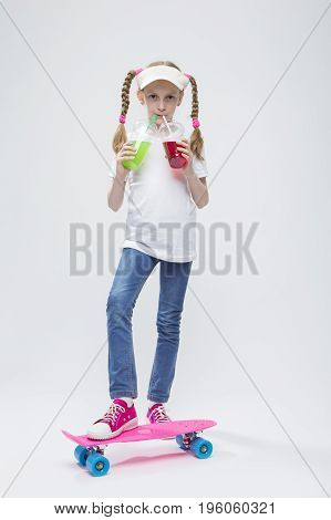 Kids Concepts. Portrait of Little Caucasian Blond Girl in Visor Posing on Pennyboard With Two Cups of Juice and Straw. Against White. Vertical Image Orientation
