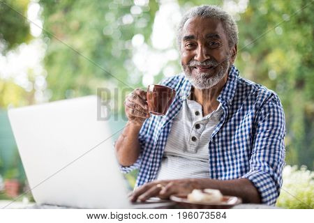 Portrait of senior man having coffee while using laptop at table in yard