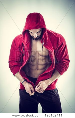 Muscular bodybuilder undressing, opening red hoodie sweater on naked muscle torso on light background